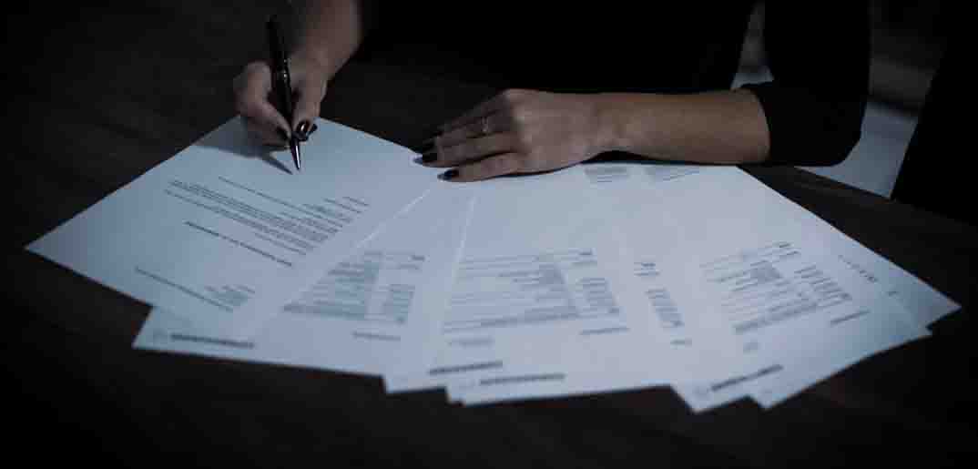 Hands signing paperwork spread across a dark table.