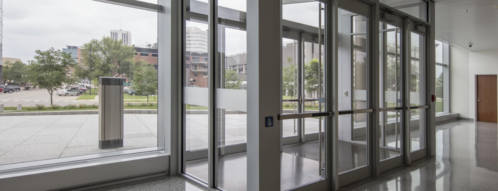 Glass entrance doors and a tall window look out from an empty lobby onto a paved plaza, young trees and parked cars.