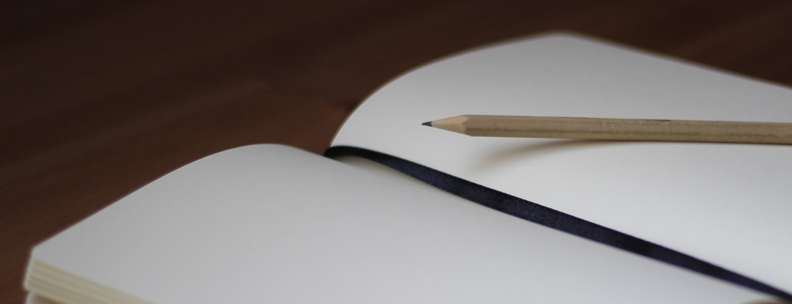 A pencil and a dark elastic band lie across the blank cream pages of an open notebook on a wooden table.